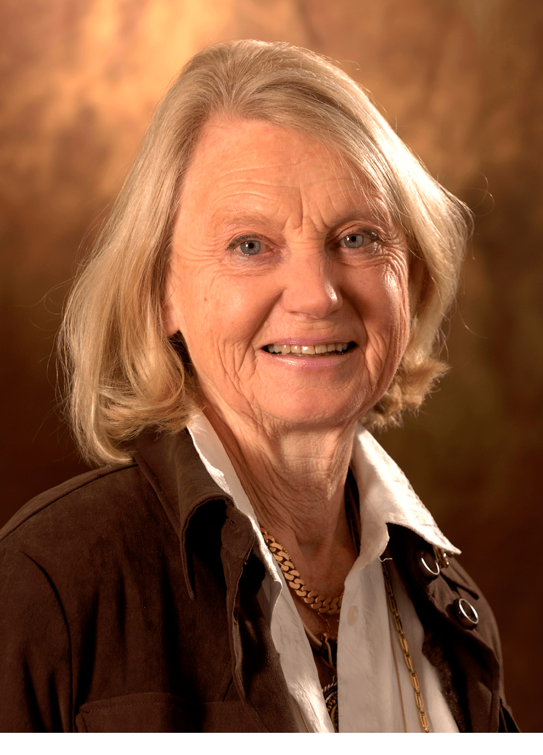 A senior with shoulder-length light hair is smiling and wearing a white collared shirt under a dark jacket with a gold chain necklace against a warm brown background. Description generated by AI