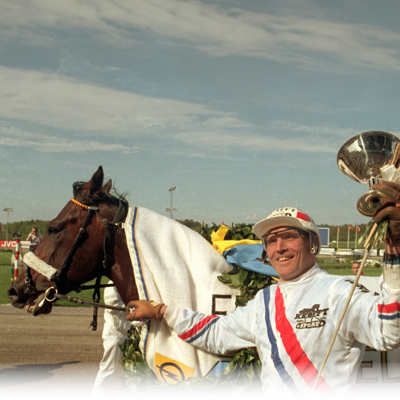 A person wearing a white racing outfit with red and blue stripes is holding the reins of a horse draped in a white blanket and adorned with a wreath, standing on a racetrack. Description generated by AI