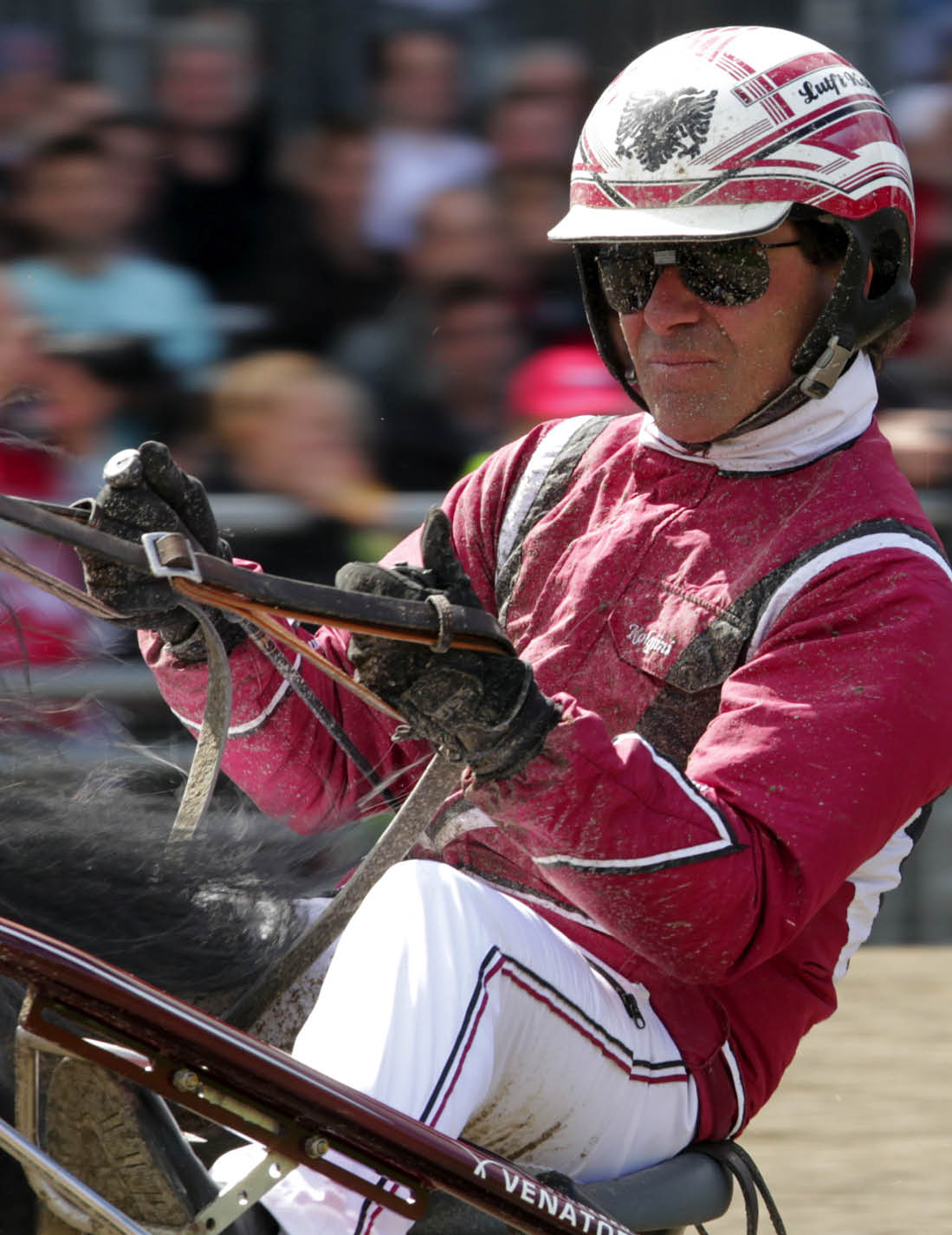 A person wearing a red and white racing outfit and helmet is holding reins while riding a horse in a competitive harness racing event. Description generated by AI