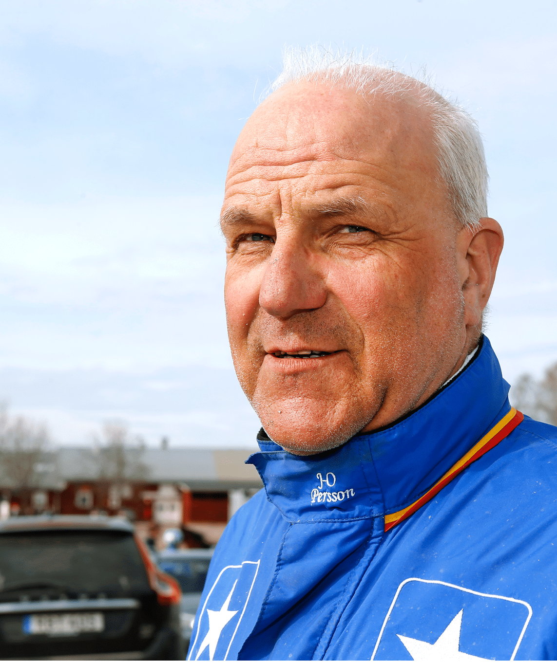 A senior person wearing a blue jacket with white star logos stands outdoors near a parked car and buildings under a cloudy sky. Description generated by AI