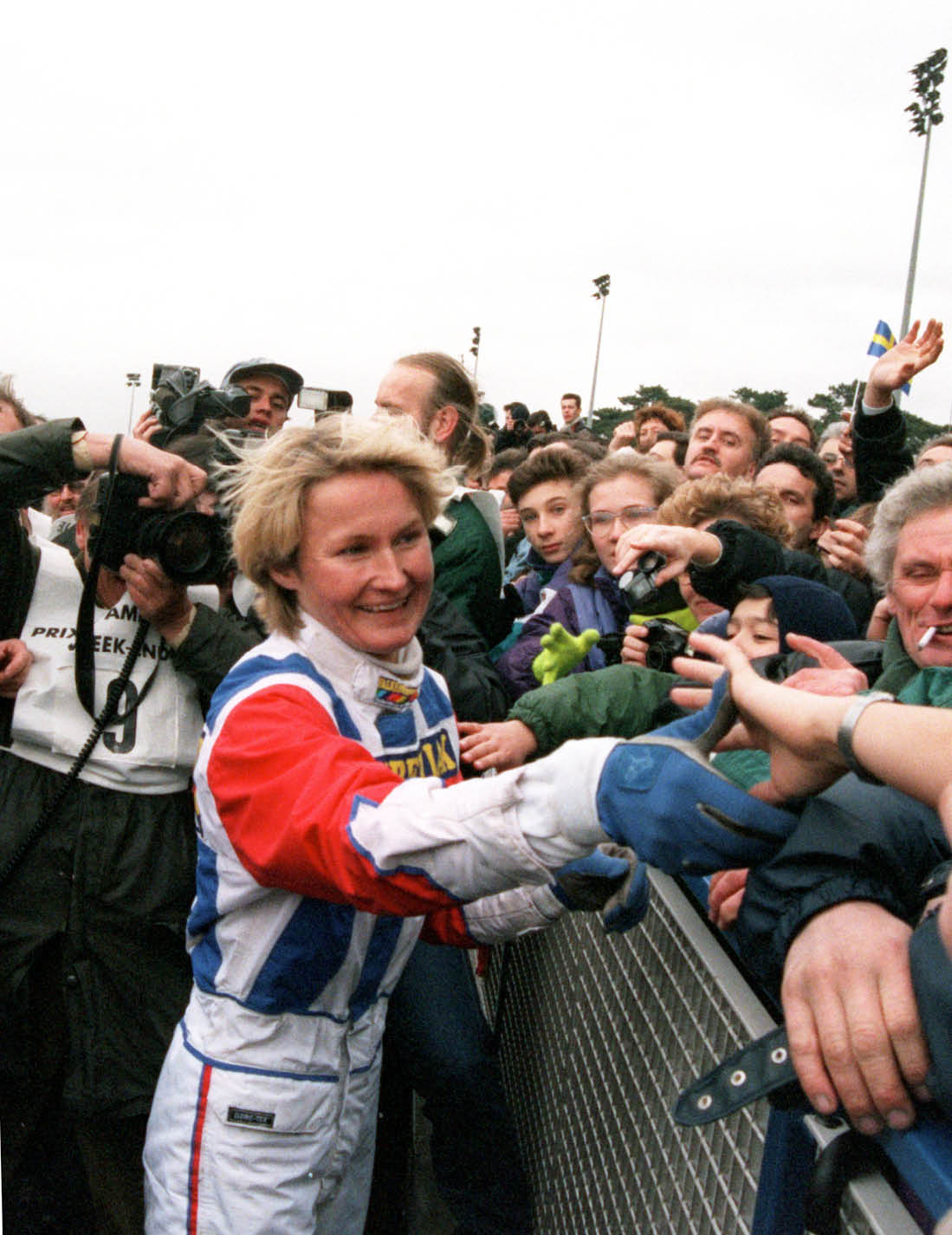 A person in a colorful racing suit is smiling and shaking hands with a crowd of people behind a barrier while photographers capture the moment. Description generated by AI