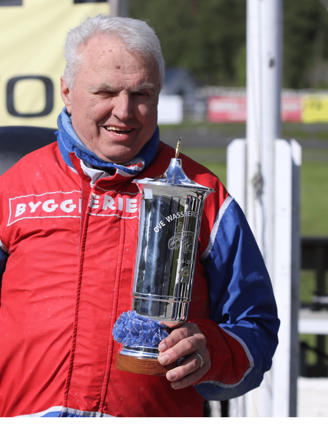 A senior wearing a red and blue racing suit is smiling and holding a silver trophy with a blue ribbon. Description generated by AI