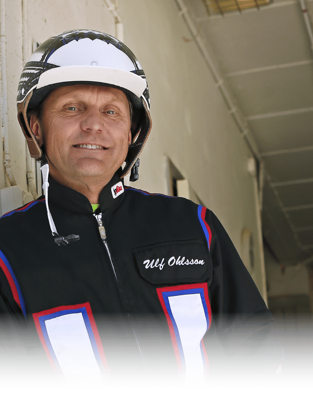 A person wearing a helmet and a black racing suit with red, blue, and white accents is smiling while standing indoors. Description generated by AI