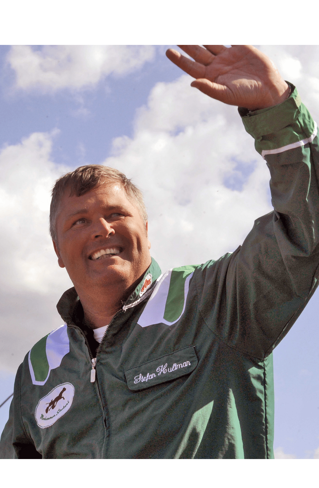A person wearing a green jacket with white and green stripes is smiling and waving against a partly cloudy sky. Description generated by AI