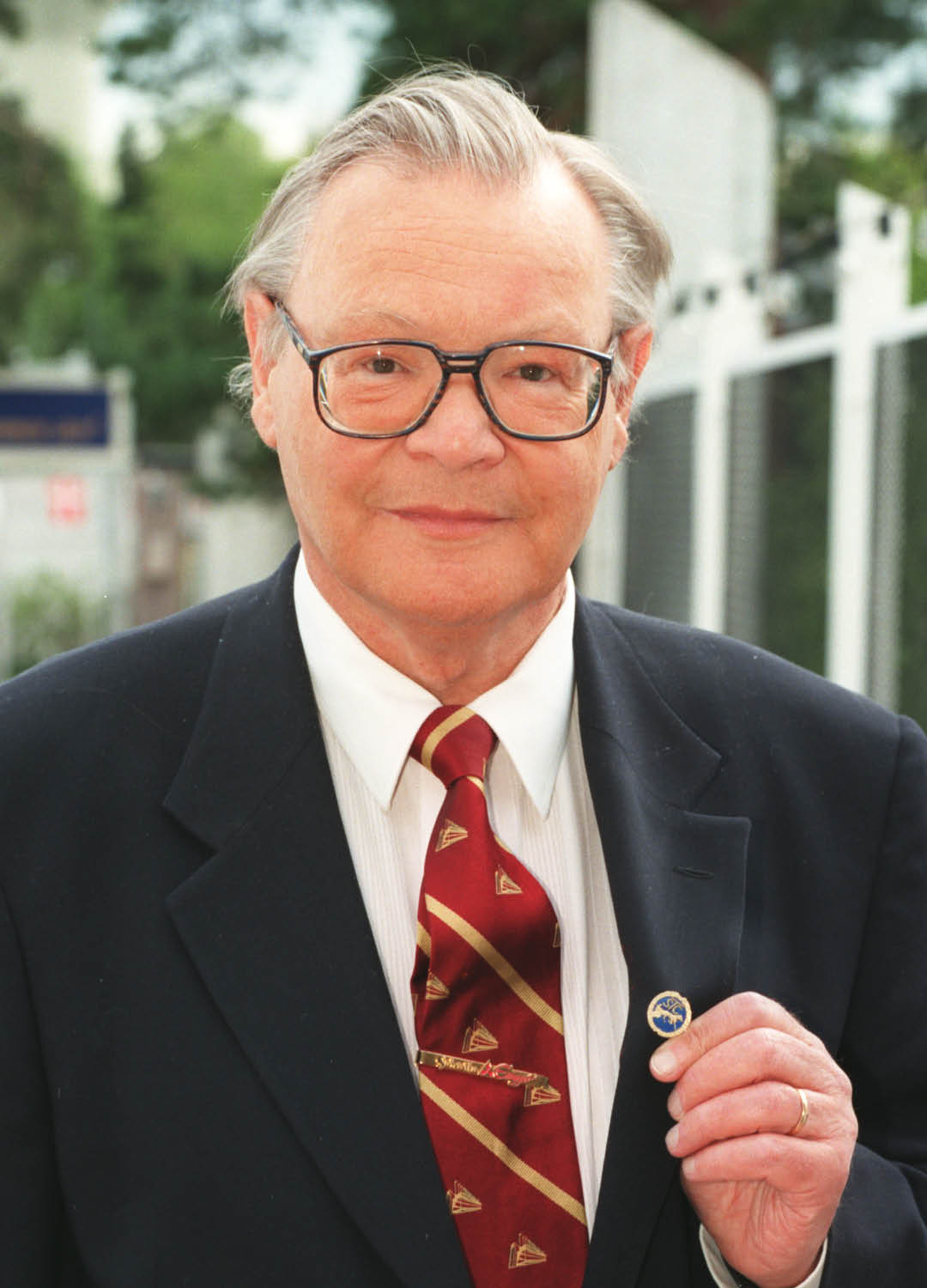 A senior person wearing glasses, a dark suit, white shirt, and patterned red tie is holding a small round pin while standing outdoors. Description generated by AI